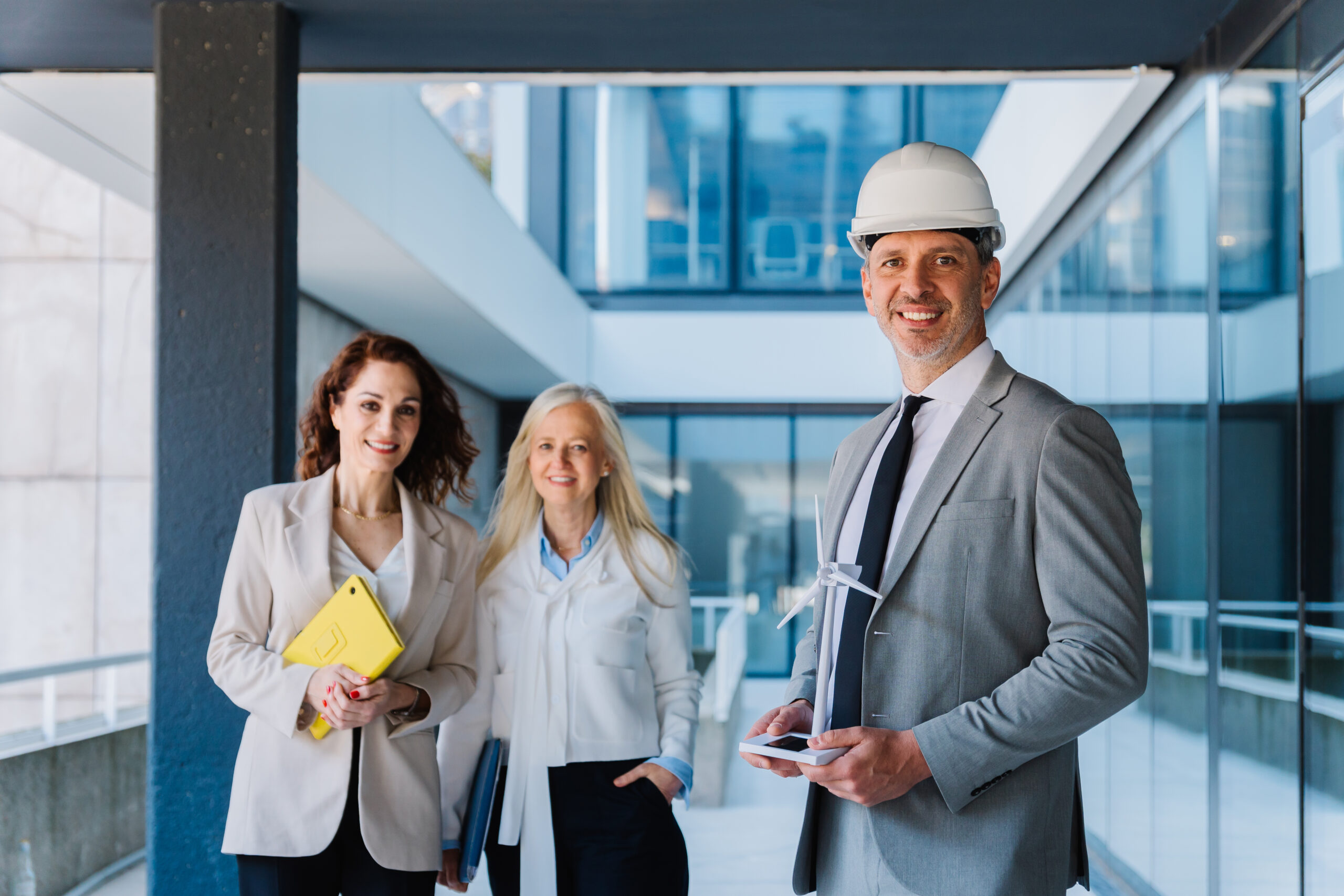 Engineer wearing a safety helmet holding a miniature wind turbine, smiling near his colleagues, businesswomen, in a modern office building
